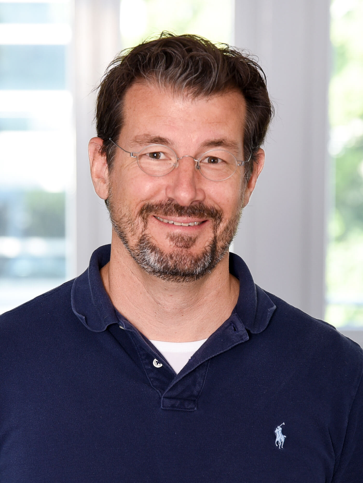 Sie sehen einen lächelnden Mann mittleren Alters mit kurzem, leicht gelockten dunklen Haar und Bart. Er trägt eine runde, fein silbern gerahmte Brille und ein dunkelblaues Poloshirt.